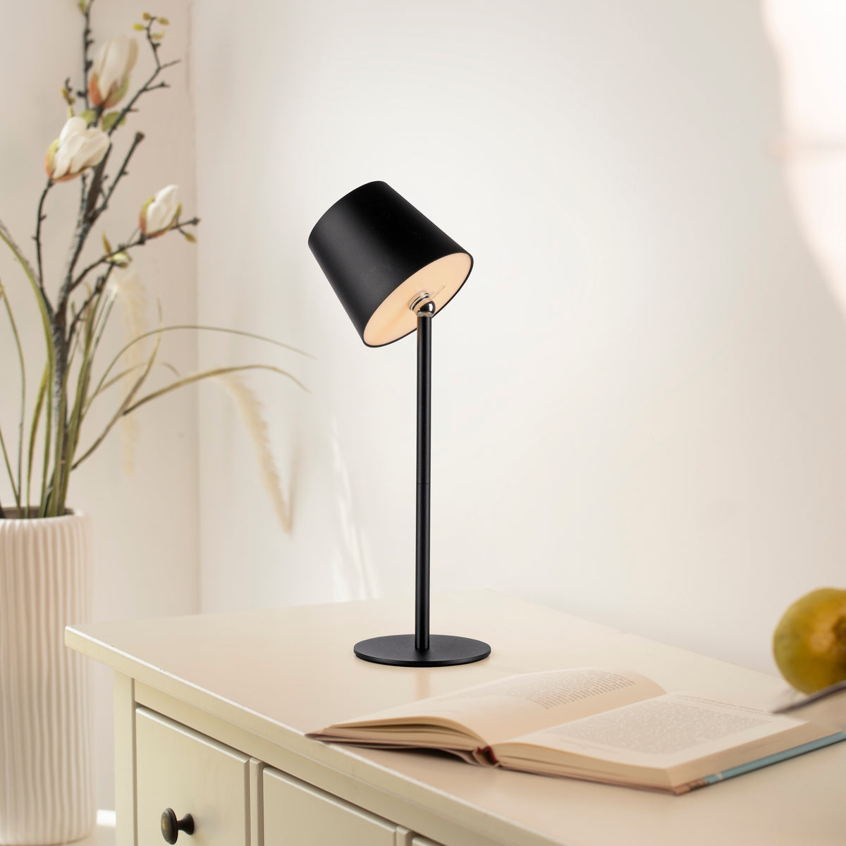 Lampe à poser noire allumée sur un meuble en bois avec livre ouvert et vase décoratif.