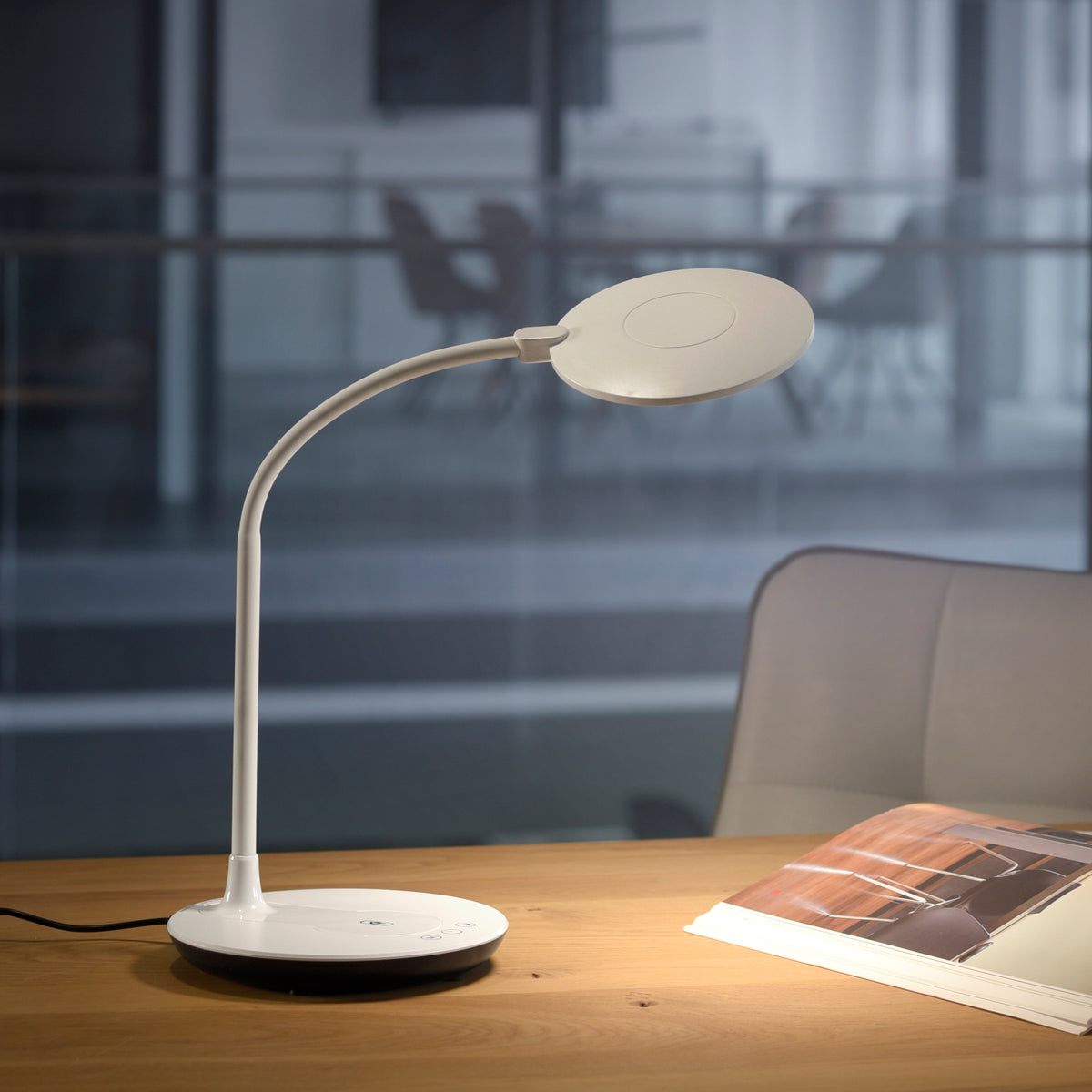 Lampe allumée posée sur un bureau en bois près d'un magazine, dans un bureau vitré.