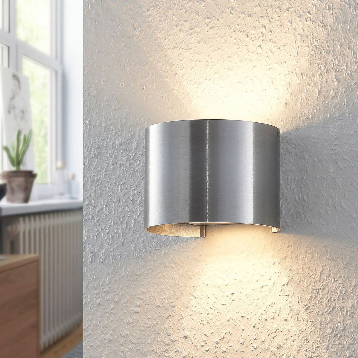Applique en aluminium brossé fixée au mur près d'une fenêtre, avec plante et radiateur en arrière-plan.