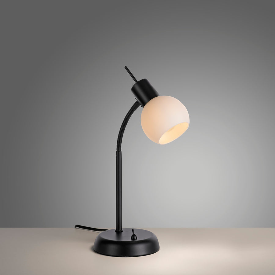 Lampe de bureau noire inclinée, abat-jour sphérique allumé, posée sur surface taupe.