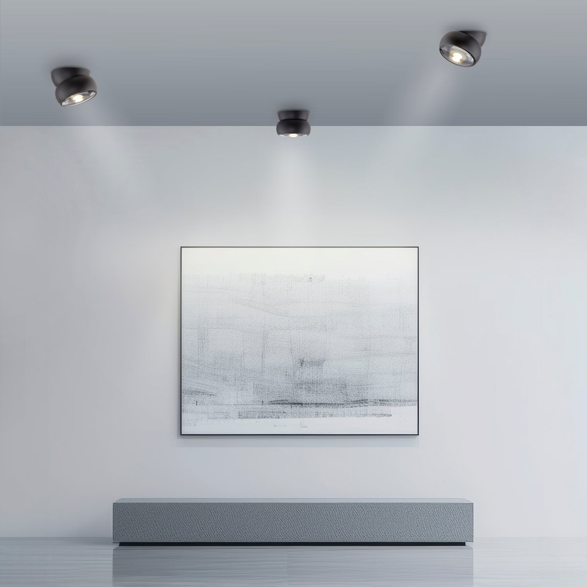 Trois plafonniers noirs orientables éclairant un mur blanc avec une œuvre encadrée au-dessus d'un meuble.