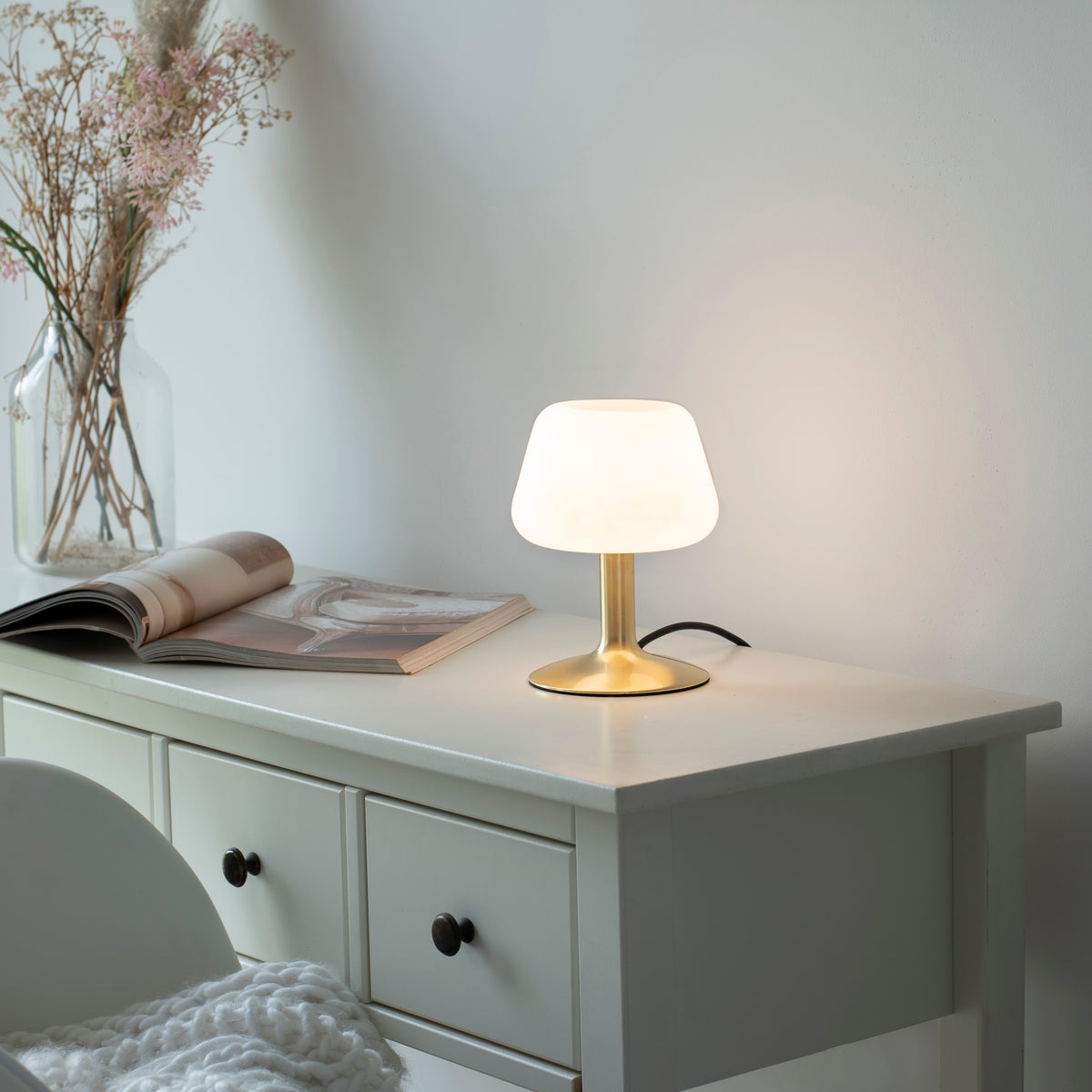 Mise en ambiance sur commode blanche, lampe allumée près d'un magazine et de fleurs séchées.