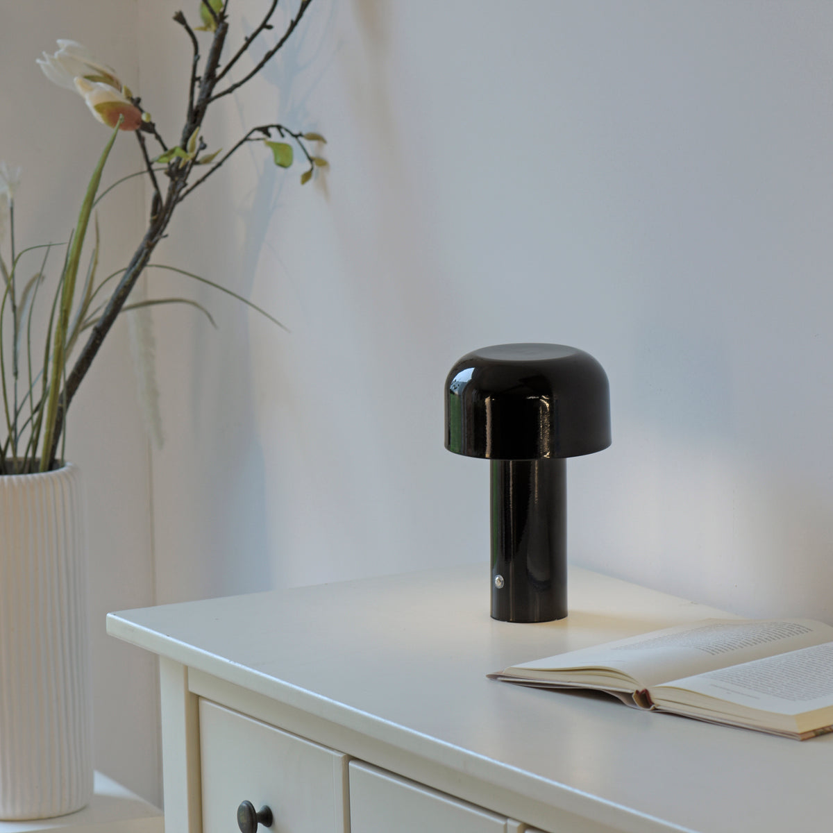 Lampe noire posée sur un meuble blanc près d'un livre ouvert et d'un vase à fleurs.