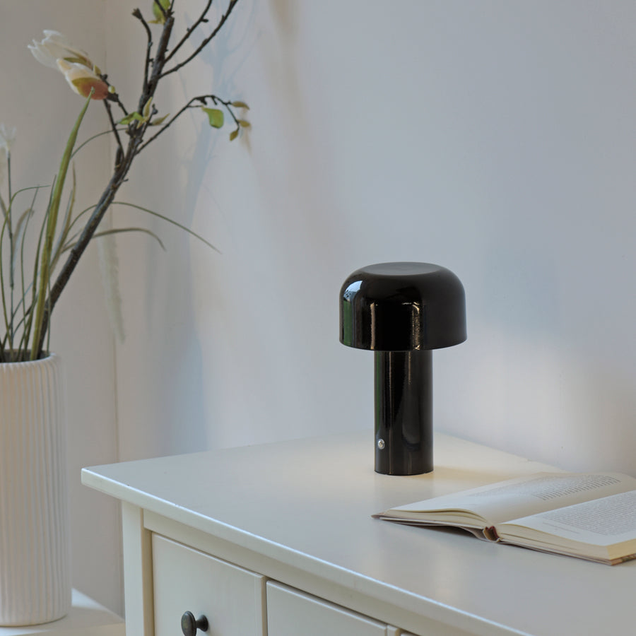 Lampe noire posée sur un meuble blanc près d'un livre ouvert et d'un vase à fleurs.