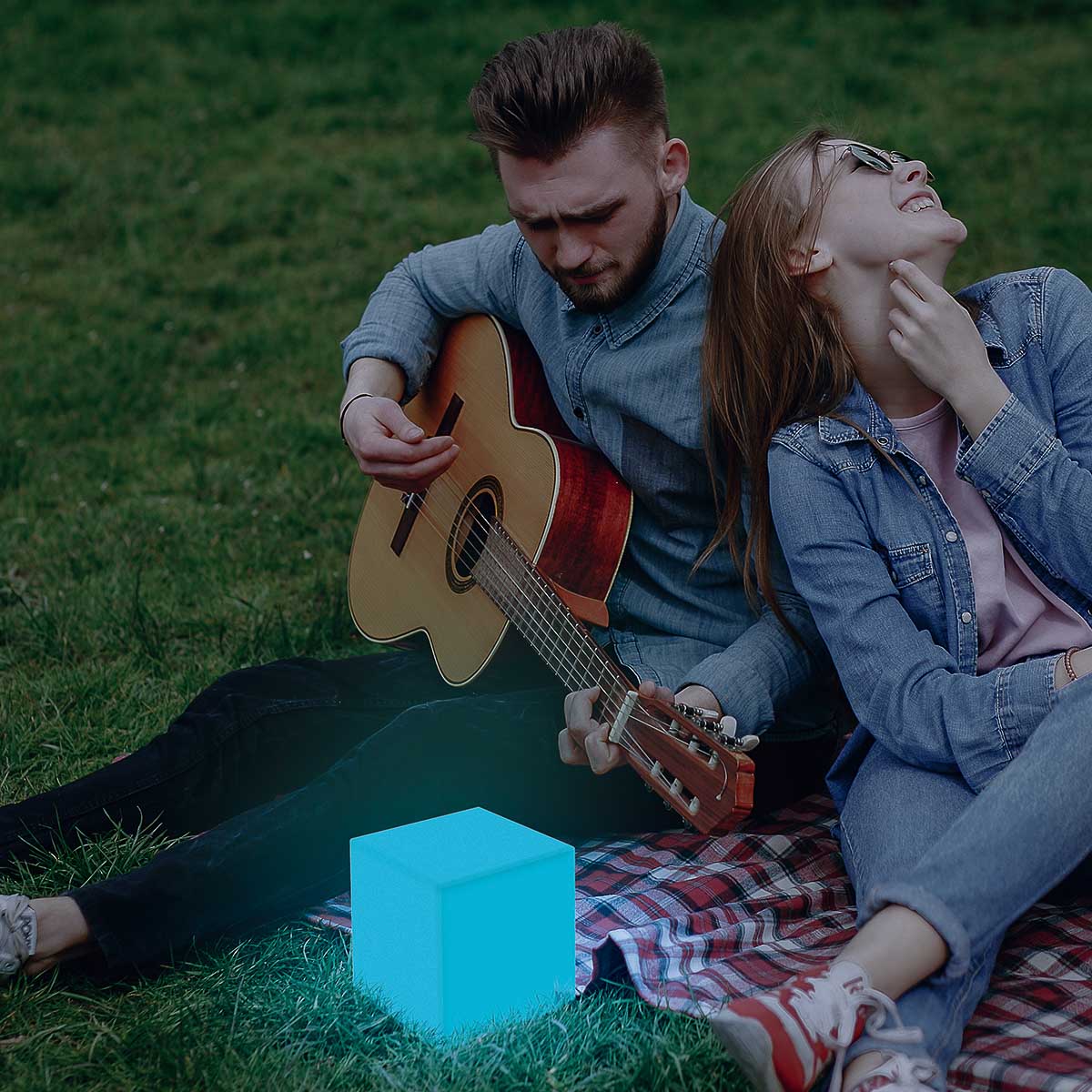Mise en ambiance : couple assis sur plaid, cube lumineux bleu sur herbe