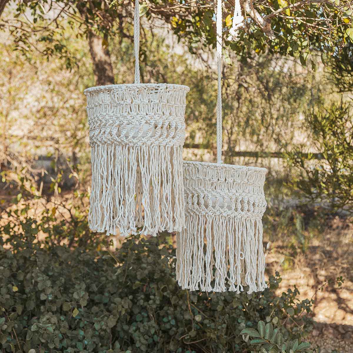 Deux abat-jours en macramé à franges suspendus dans un jardin ensoleillé.