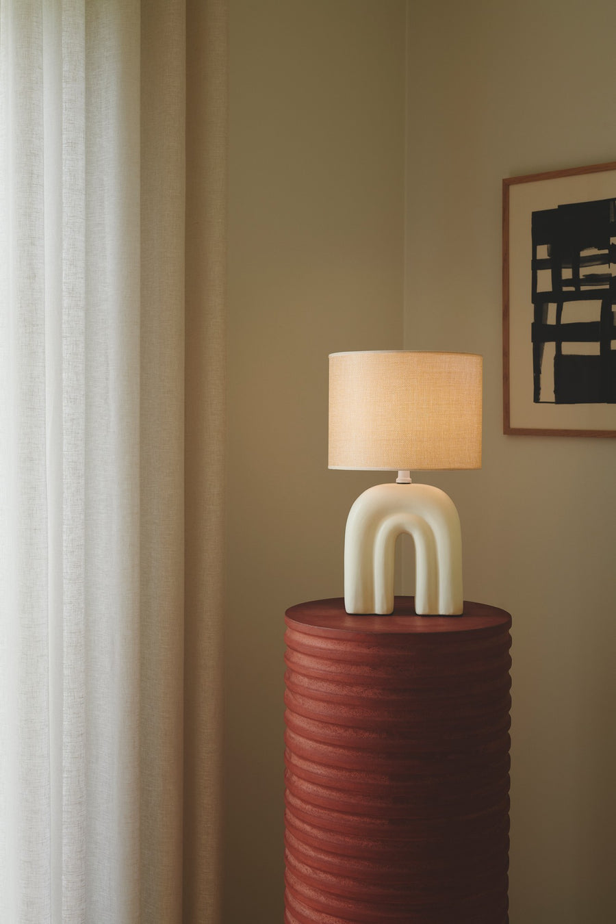 Lampe allumée posée sur un tabouret rouge, devant rideaux clairs et cadre mural