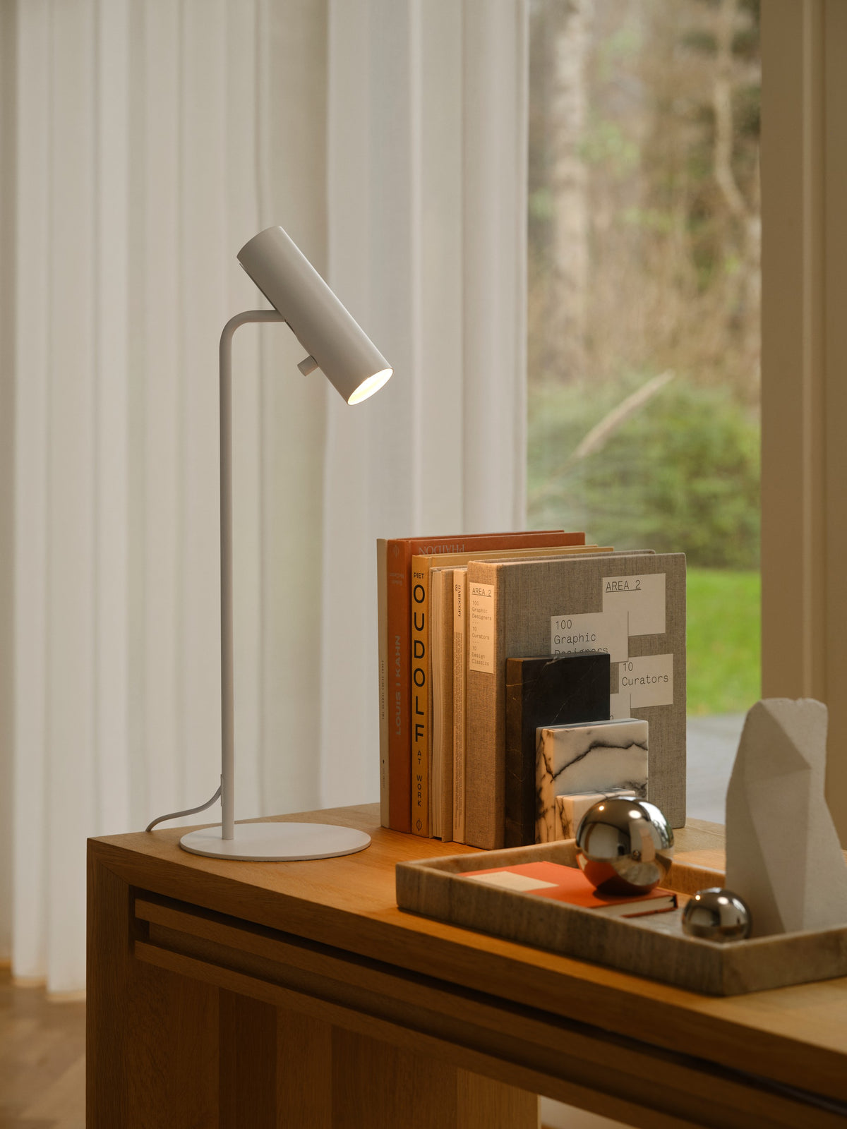 Mise en ambiance : lampe blanche allumée posée sur un meuble en bois devant une fenêtre.