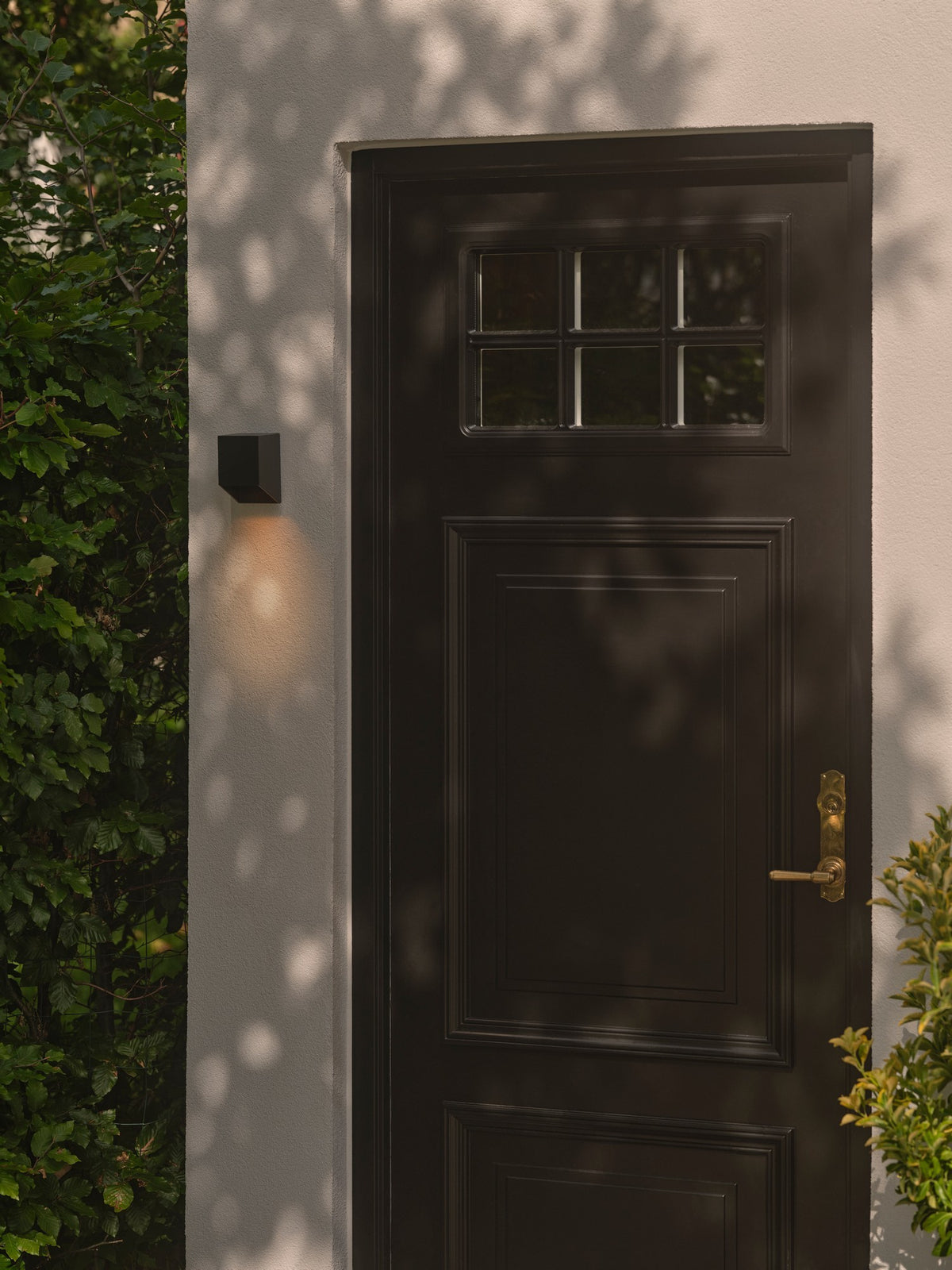 Applique noire installée sur un mur extérieur près d'une porte, éclairant le mur vers le bas.