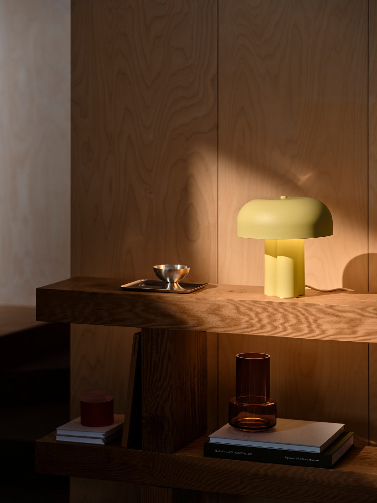 Mise en ambiance sur une étagère en bois, lampe allumée près d'une coupelle métallique.