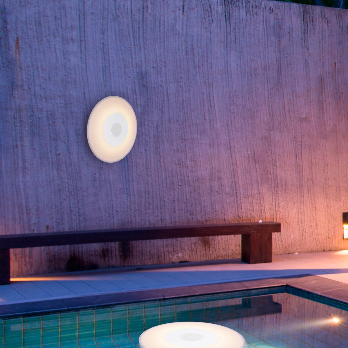 Enceinte blanche ronde fixée au mur en béton, éclairant une terrasse au bord de la piscine.