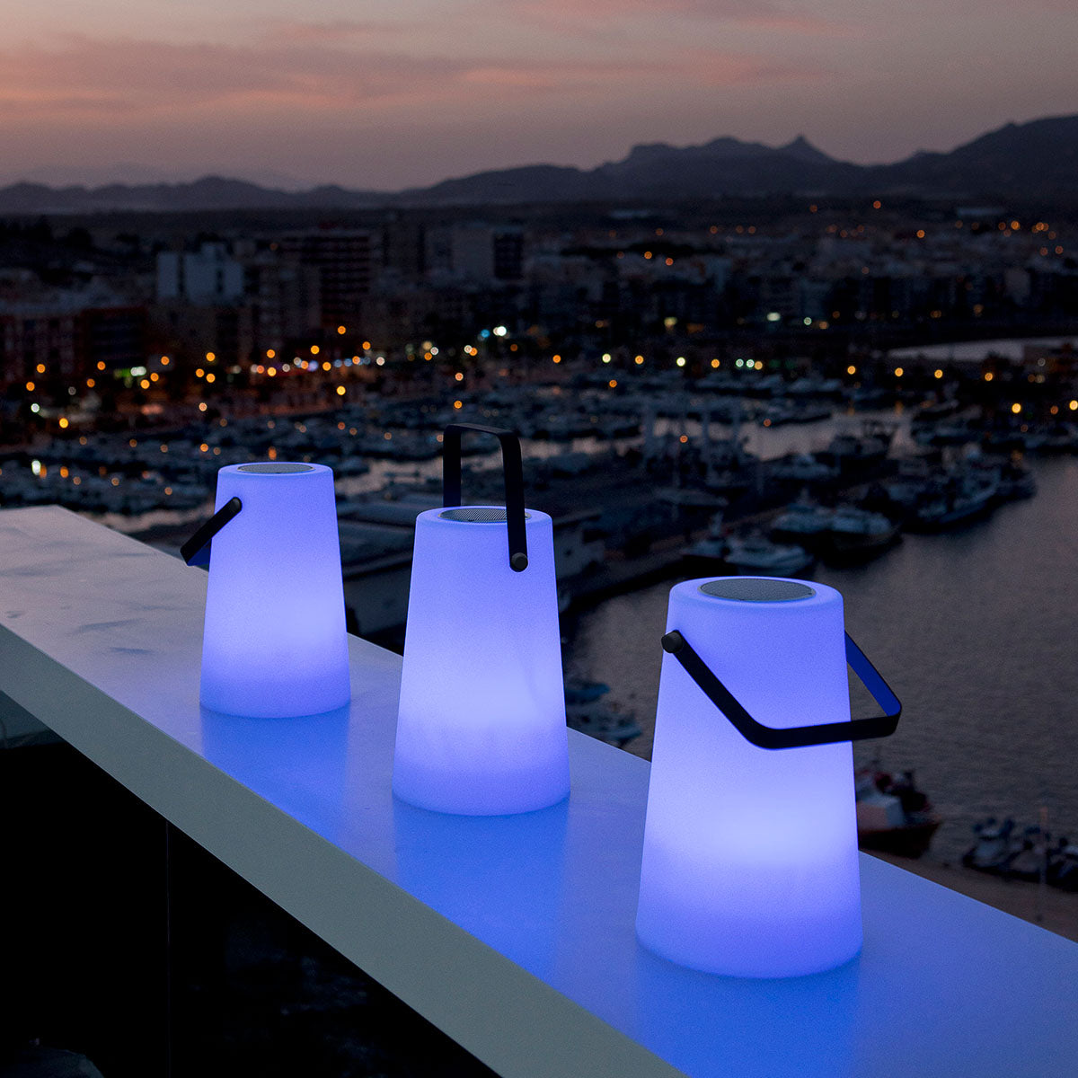 Trois lampes nomades bleues alignées sur un garde-corps face au port au crépuscule
