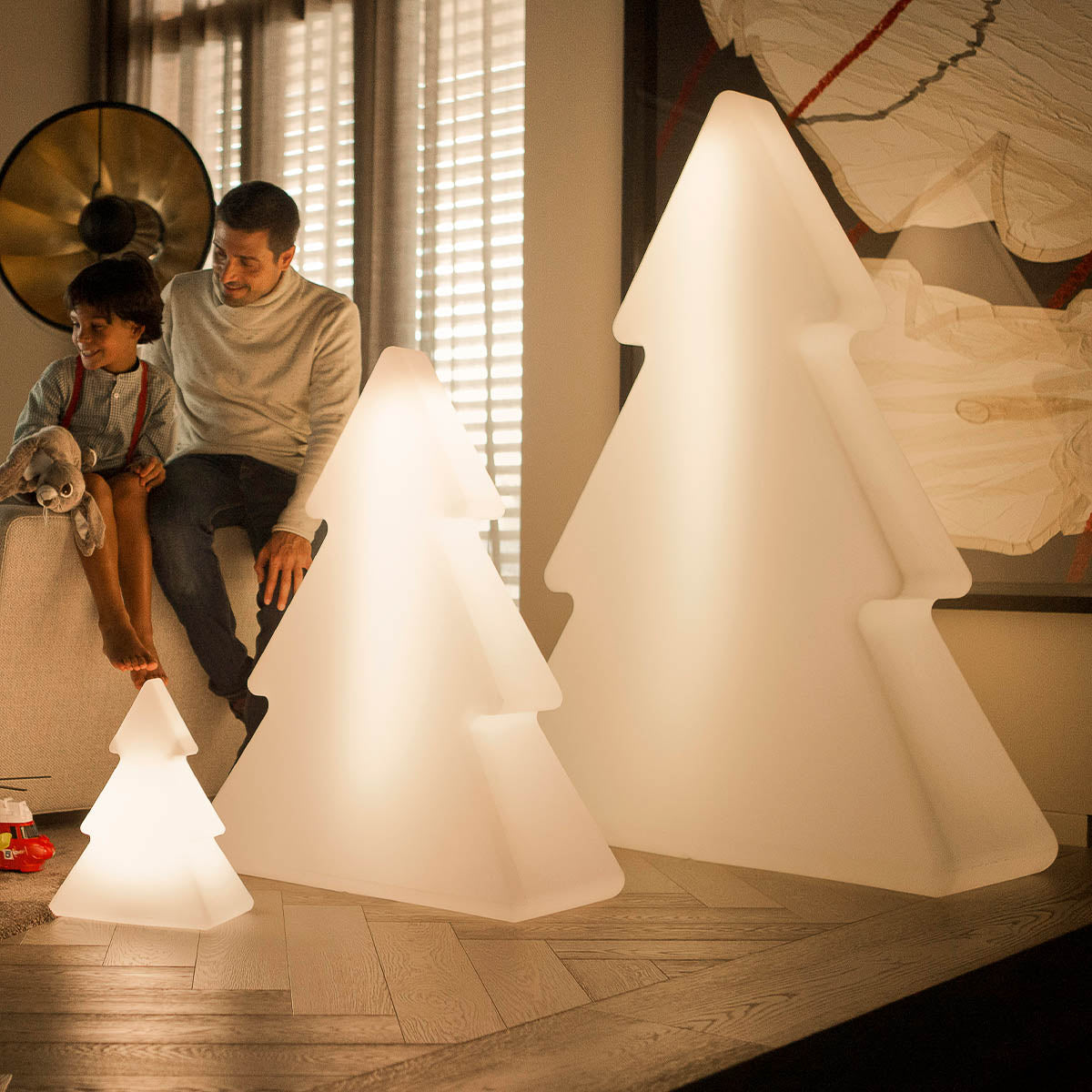 Trois sapins lumineux au sol, enfant et adulte souriants assis sur un canapé beige