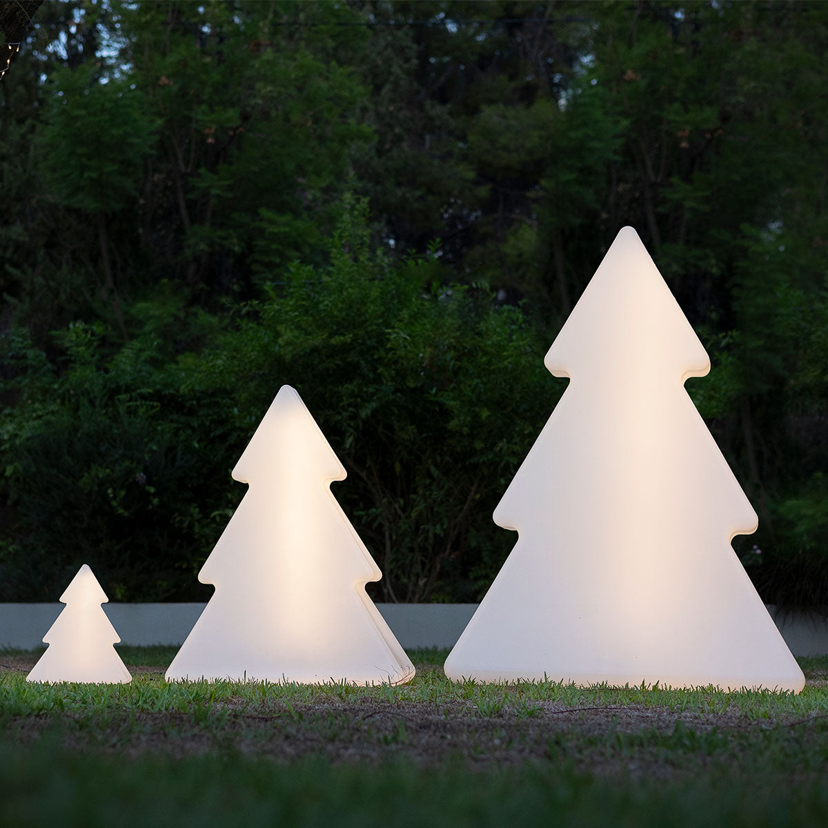Trois sapins lumineux blancs de tailles différentes alignés sur la pelouse devant des arbres