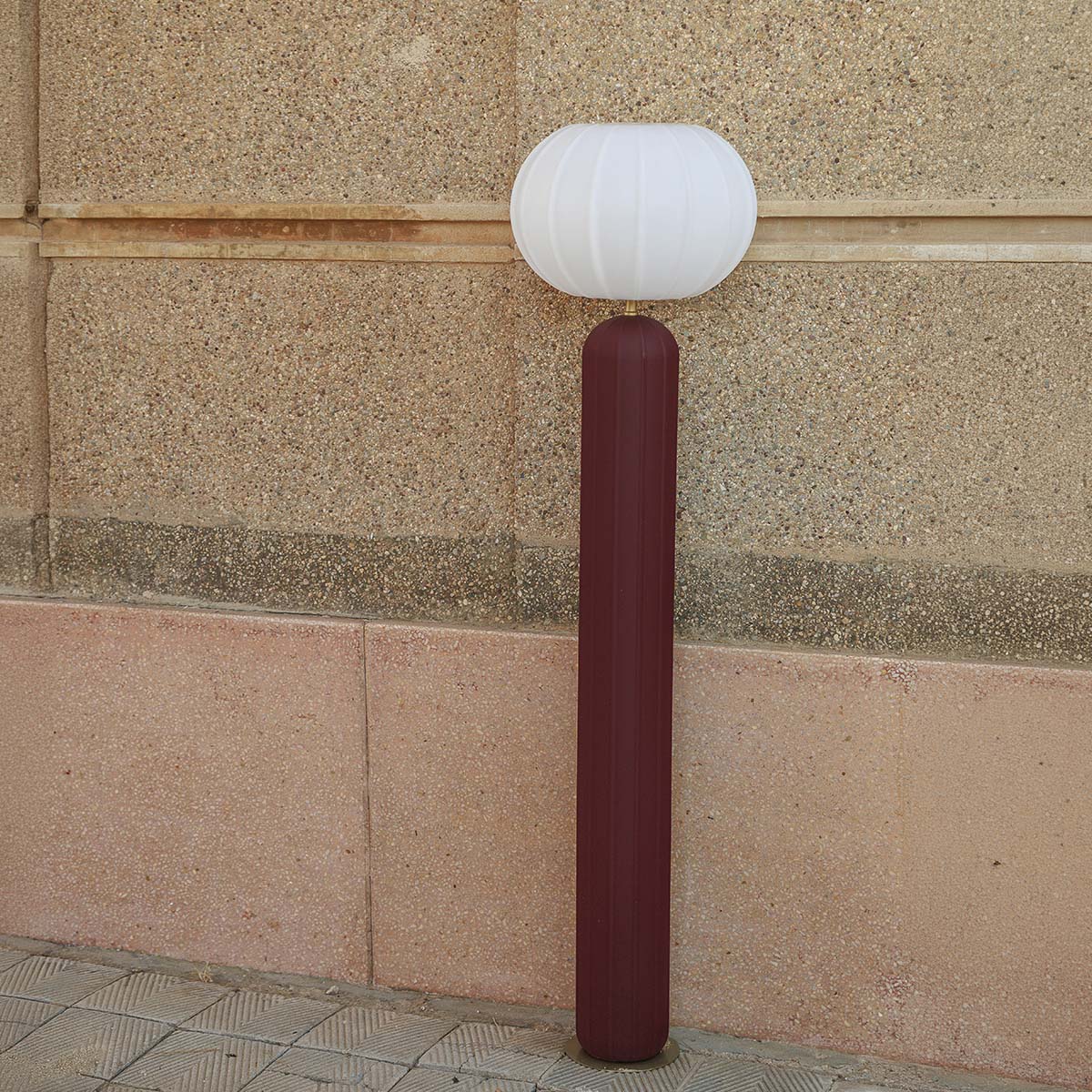Vue de face du lampadaire bordeaux devant un mur de pierre en plein jour.