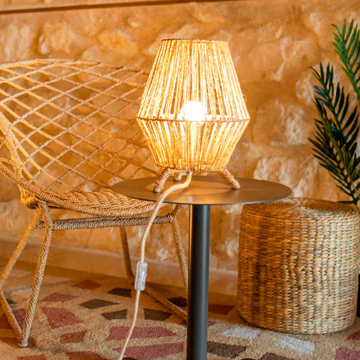 Lampe allumée sur une table d'appoint noire, fauteuil en rotin et mur en pierre.