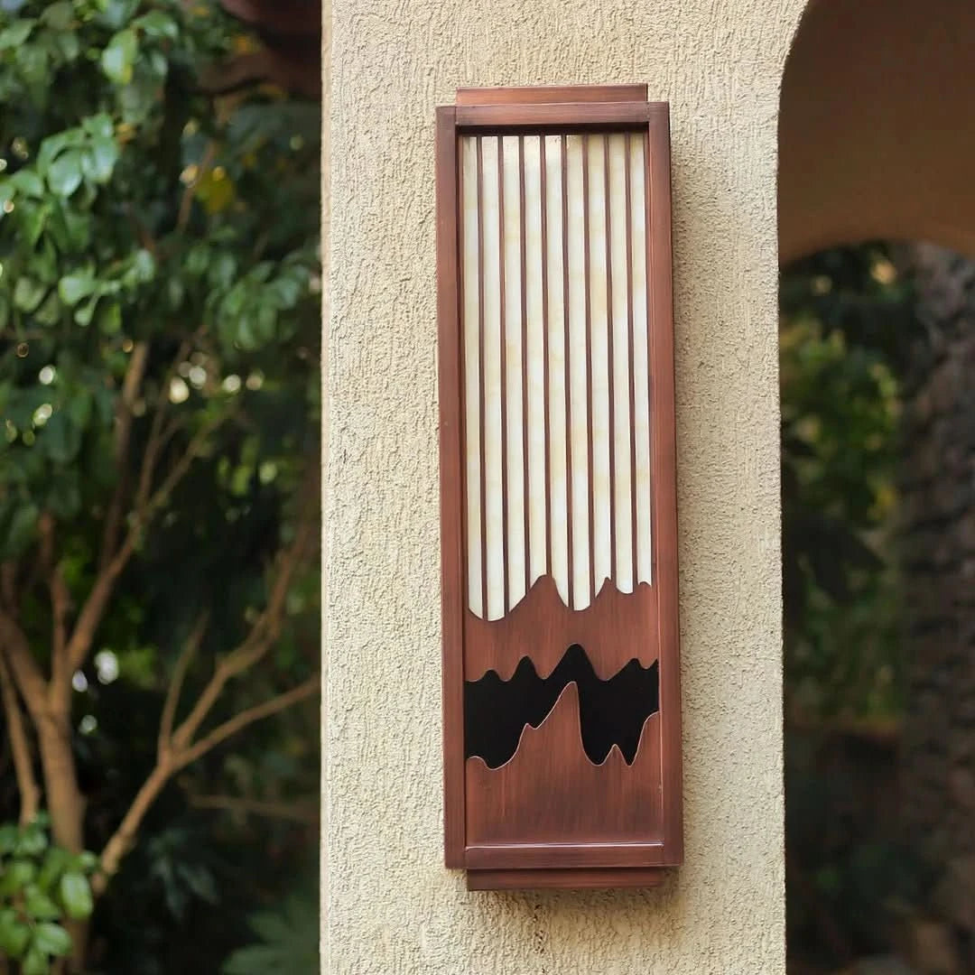Vue de l'applique murale en bois installée sur un mur extérieur crème, entourée de feuillage.