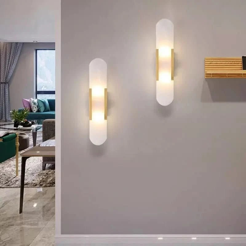 Deux appliques dorées allumées sur mur gris, salon visible à gauche