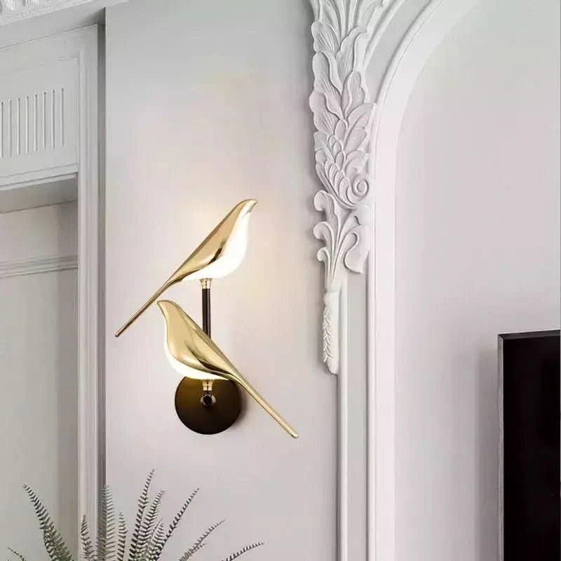 Applique murale dorée à deux oiseaux lumineux sur mur blanc mouluré, décor végétal.