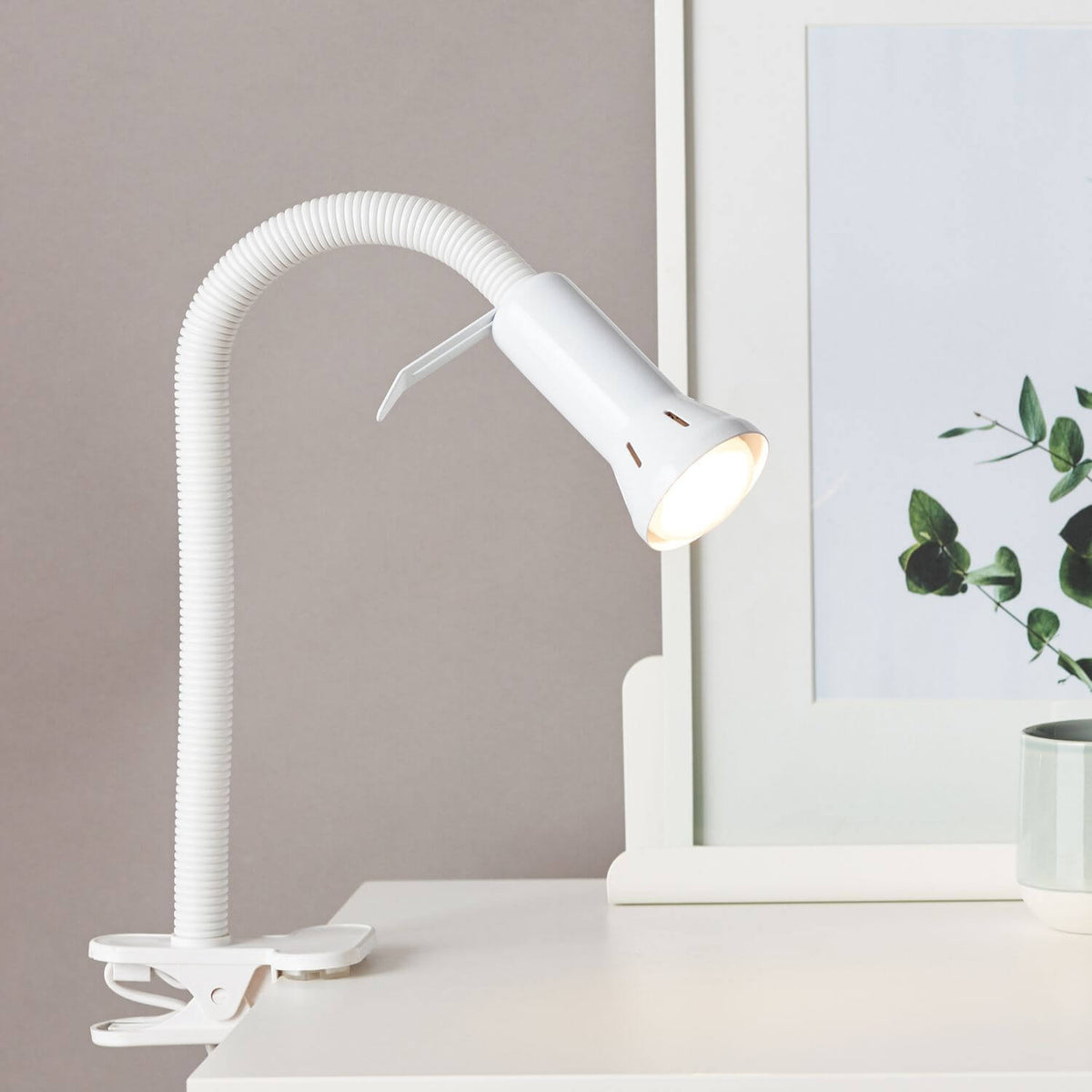 Lampe blanche allumée fixée par pince sur un bureau, éclairant une plante et une tasse.