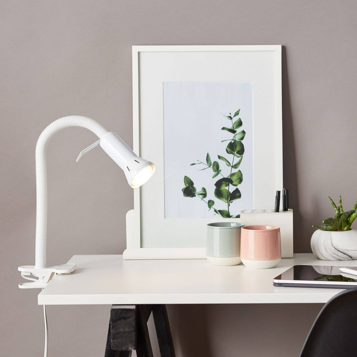Lampe de bureau blanche installée sur une table, avec cadre, tasses et plante grasse.