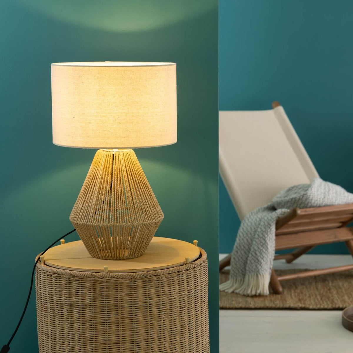 Mise en ambiance, lampe allumée sur une table d'appoint en rotin devant un mur vert.