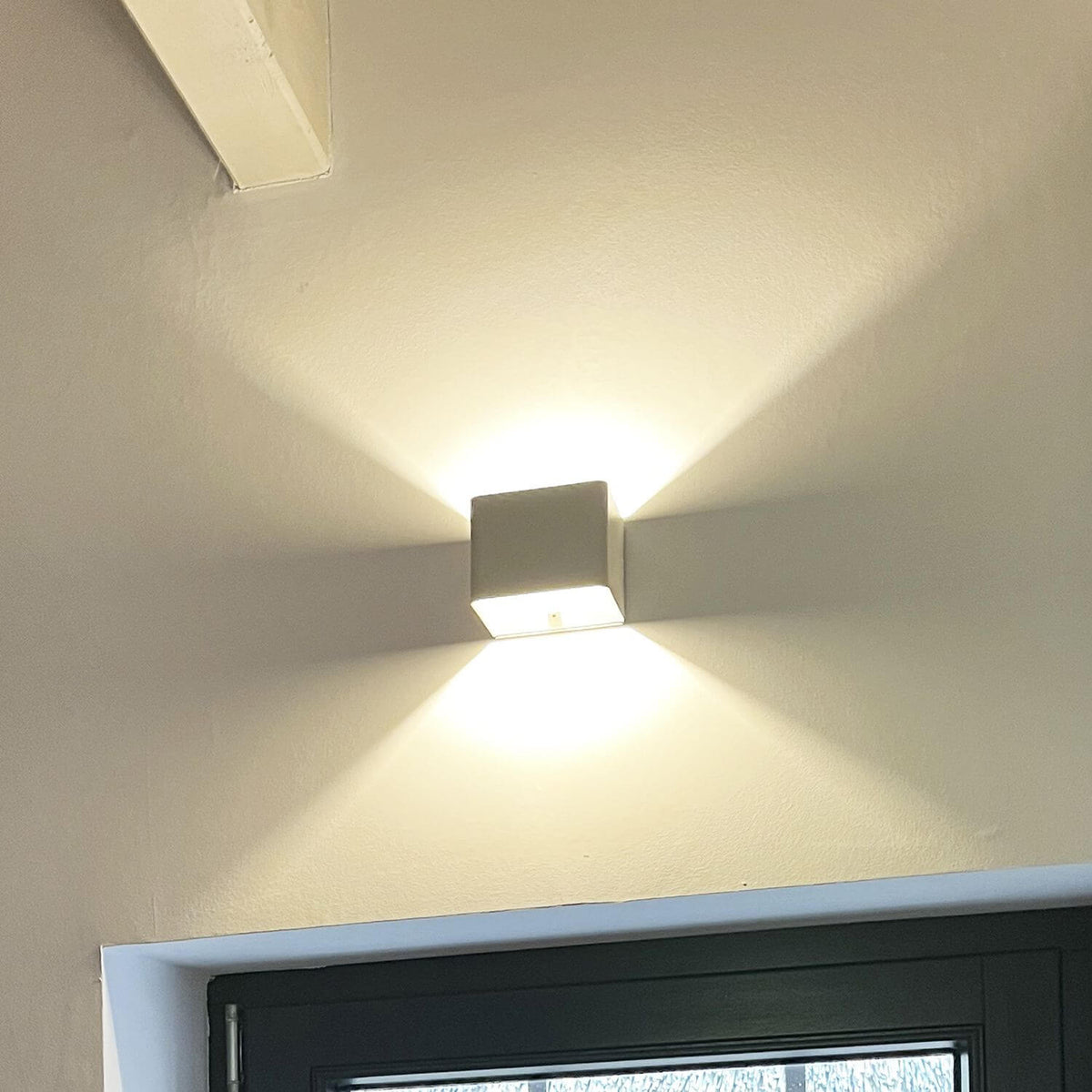 Luminaire mural blanc installé près d'une porte, lumière chaude diffusée vers le haut et le bas.