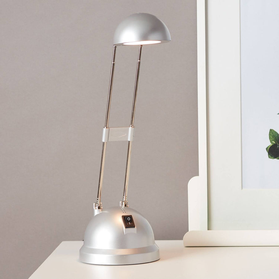 Lampe de bureau allumée posée sur une table, éclairant un cadre blanc et une plante.