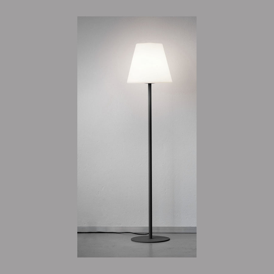 Lampadaire entier de face devant un mur clair, abat-jour conique allumé