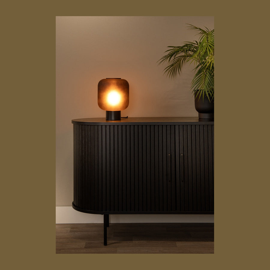 Lampe allumée posée sur un buffet noir à portes striées, plante verte à droite