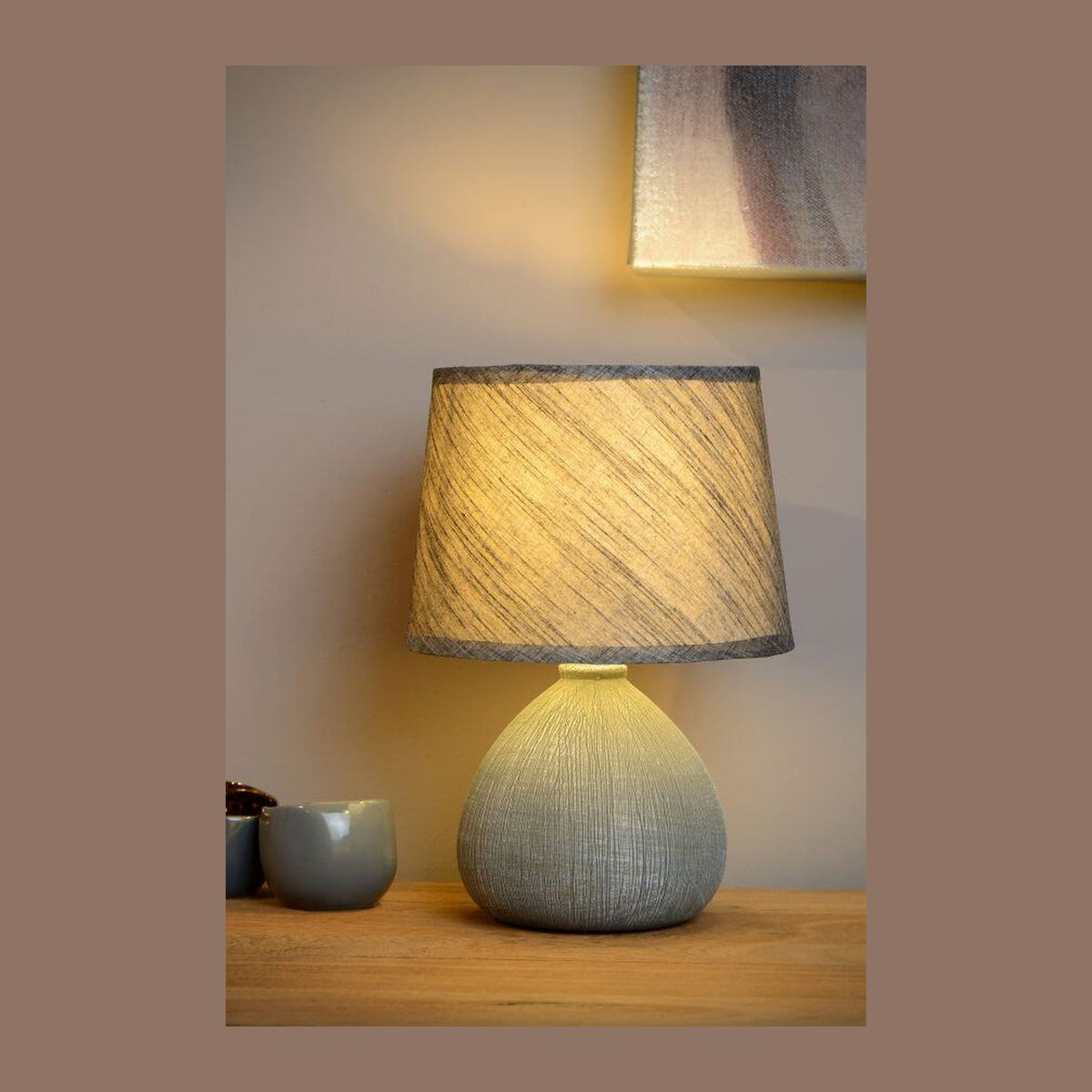 Lampe à poser allumée sur un buffet en bois, avec tasses et tableau en arrière-plan.