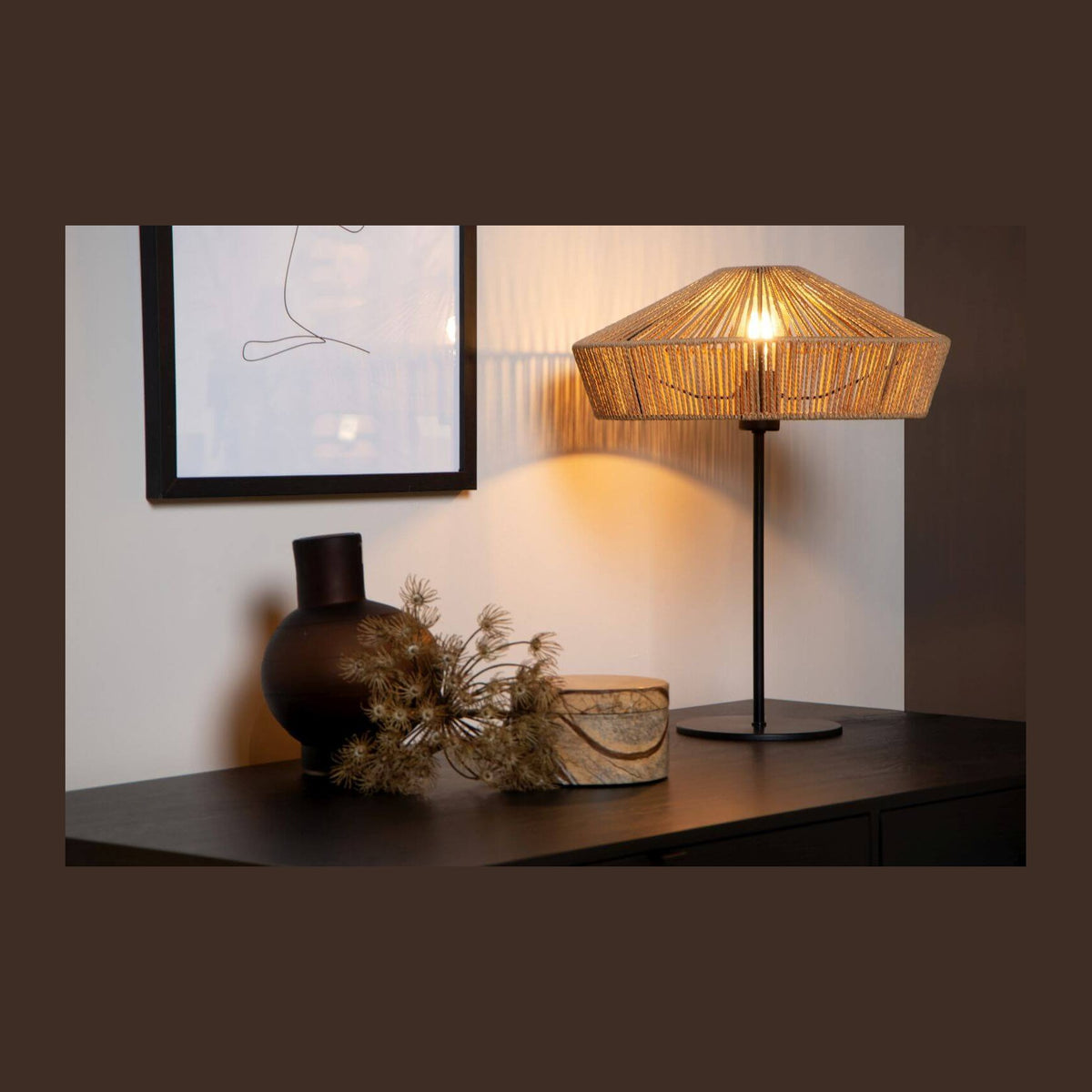 Lampe à poser allumée sur un buffet sombre, cadre et vase en arrière-plan