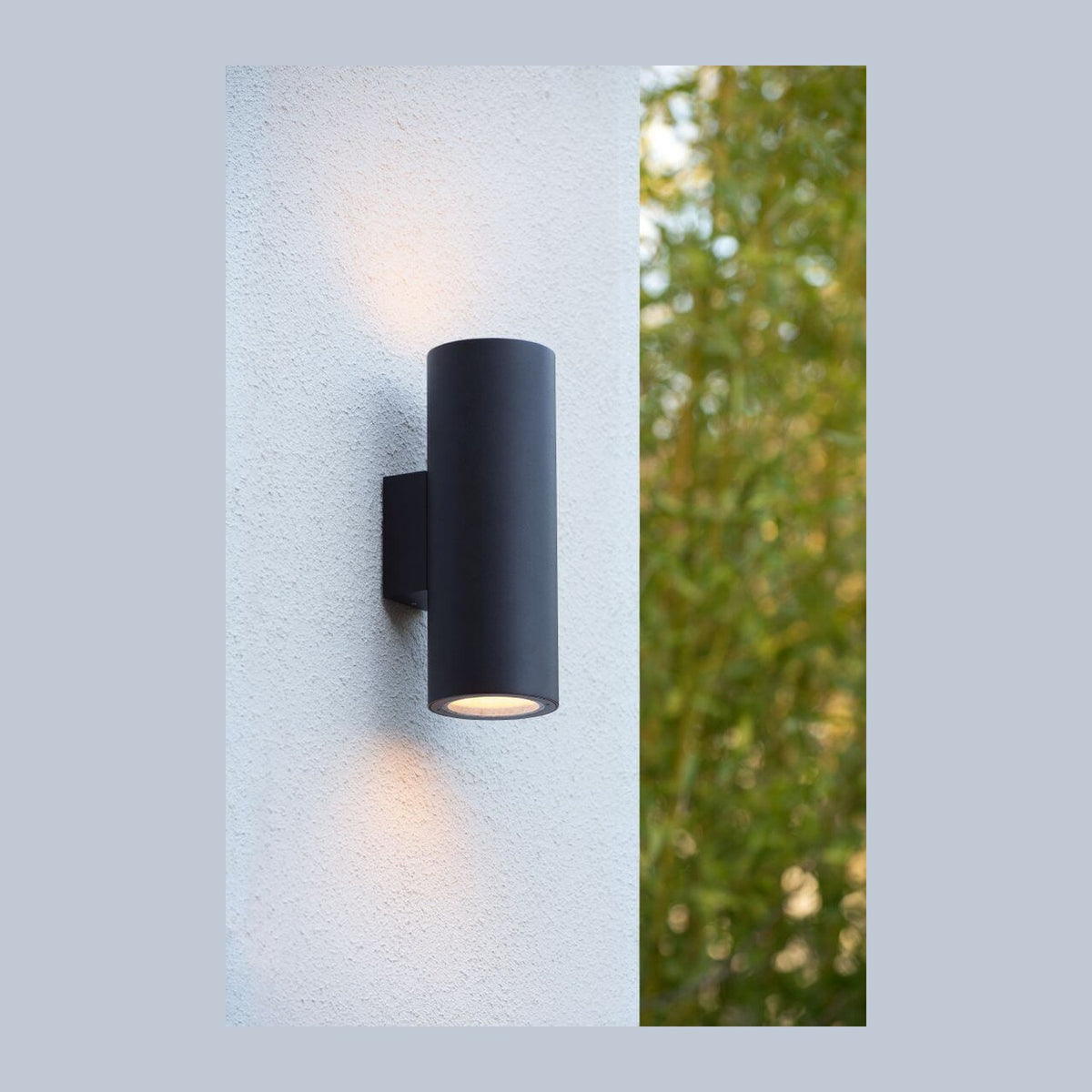Applique murale noire cylindrique sur mur blanc, feuillage flou en arrière-plan