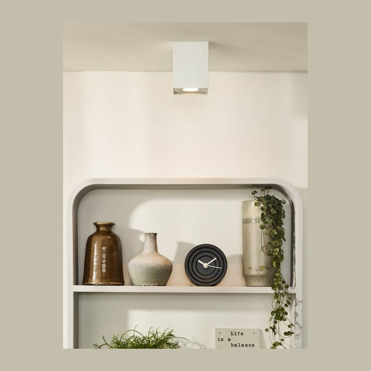Mise en ambiance : plafonnier cube blanc installé au plafond, au-dessus d’une étagère avec vases et plante.