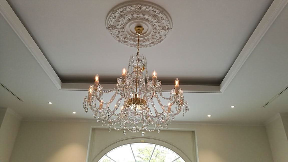 Lustre suspendu au plafond sous une rosace décorative, dans une pièce avec fenêtre cintrée.