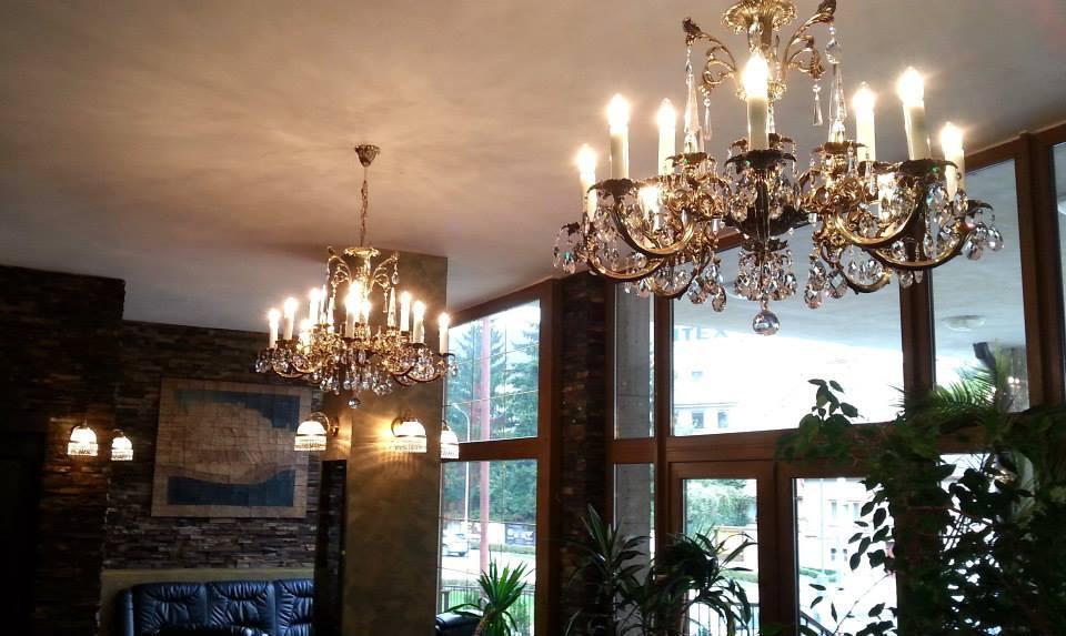 Deux lustres dorés suspendus au plafond d'un salon éclairé, avec grandes baies vitrées.