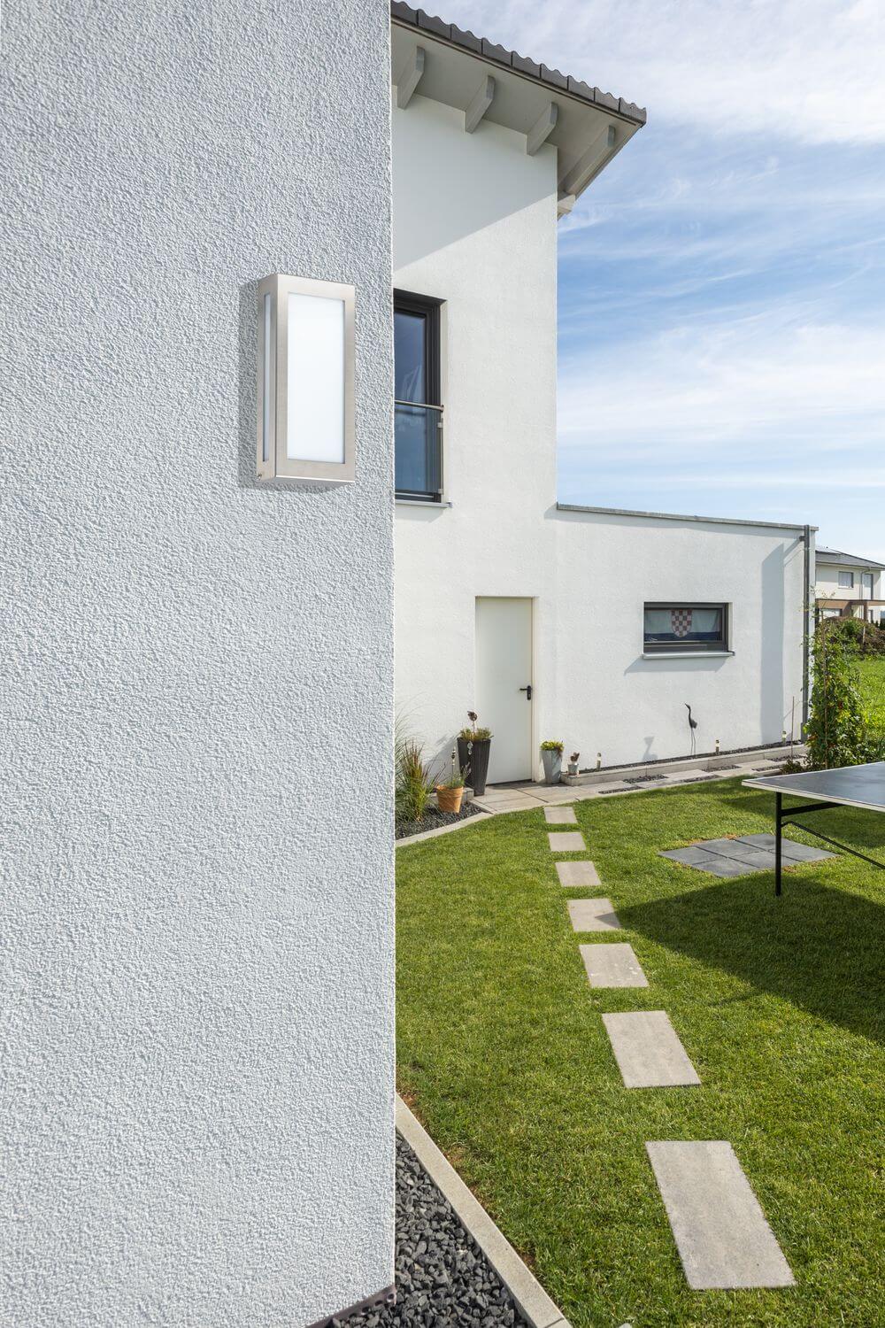 Mise en ambiance : applique allumée sur mur crépi blanc d'une maison.