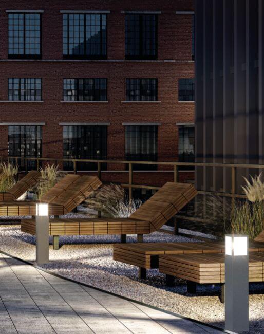 Mise en ambiance sur terrasse, deux bornes éclairent des transats devant un bâtiment.