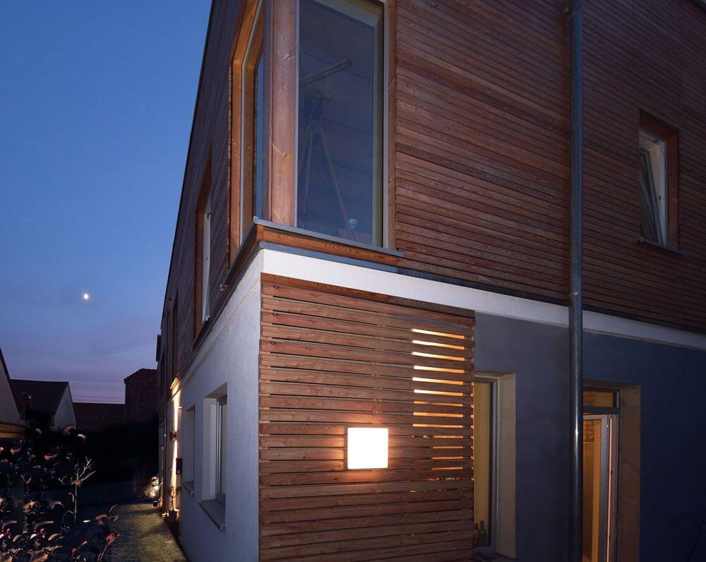 Mise en ambiance nocturne : luminaire carré allumé sur une façade en bois extérieure.