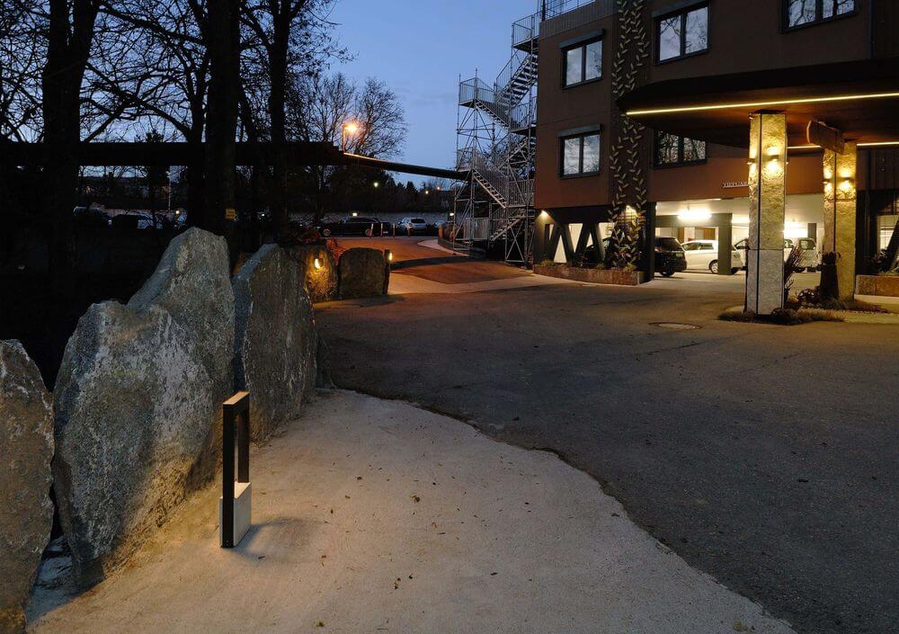 Mise en ambiance nocturne : le luminaire éclaire une allée près de rochers et d’un bâtiment.
