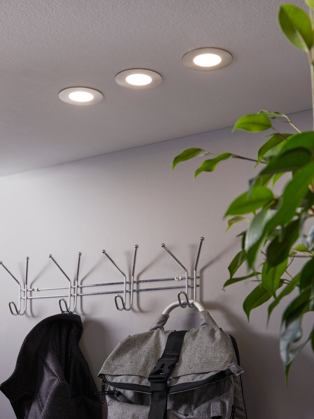 Mise en ambiance : trois spots encastrés allumés au plafond, portemanteau, sac et plante.