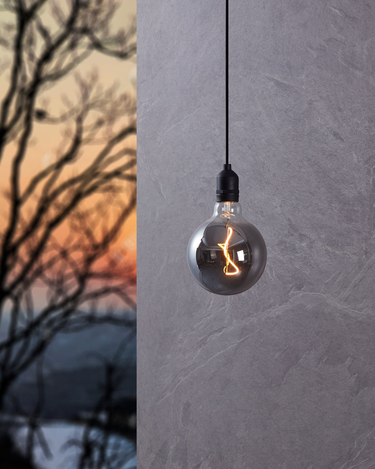 Suspension noire fixée devant un mur gris, ampoule transparente allumée, branches floues au crépuscule.