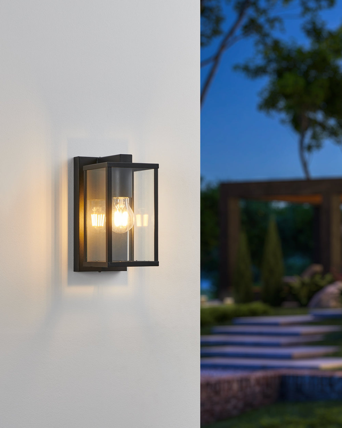 Mise en ambiance de nuit : applique allumée sur mur blanc, halo chaud, jardin et marches en arrière-plan