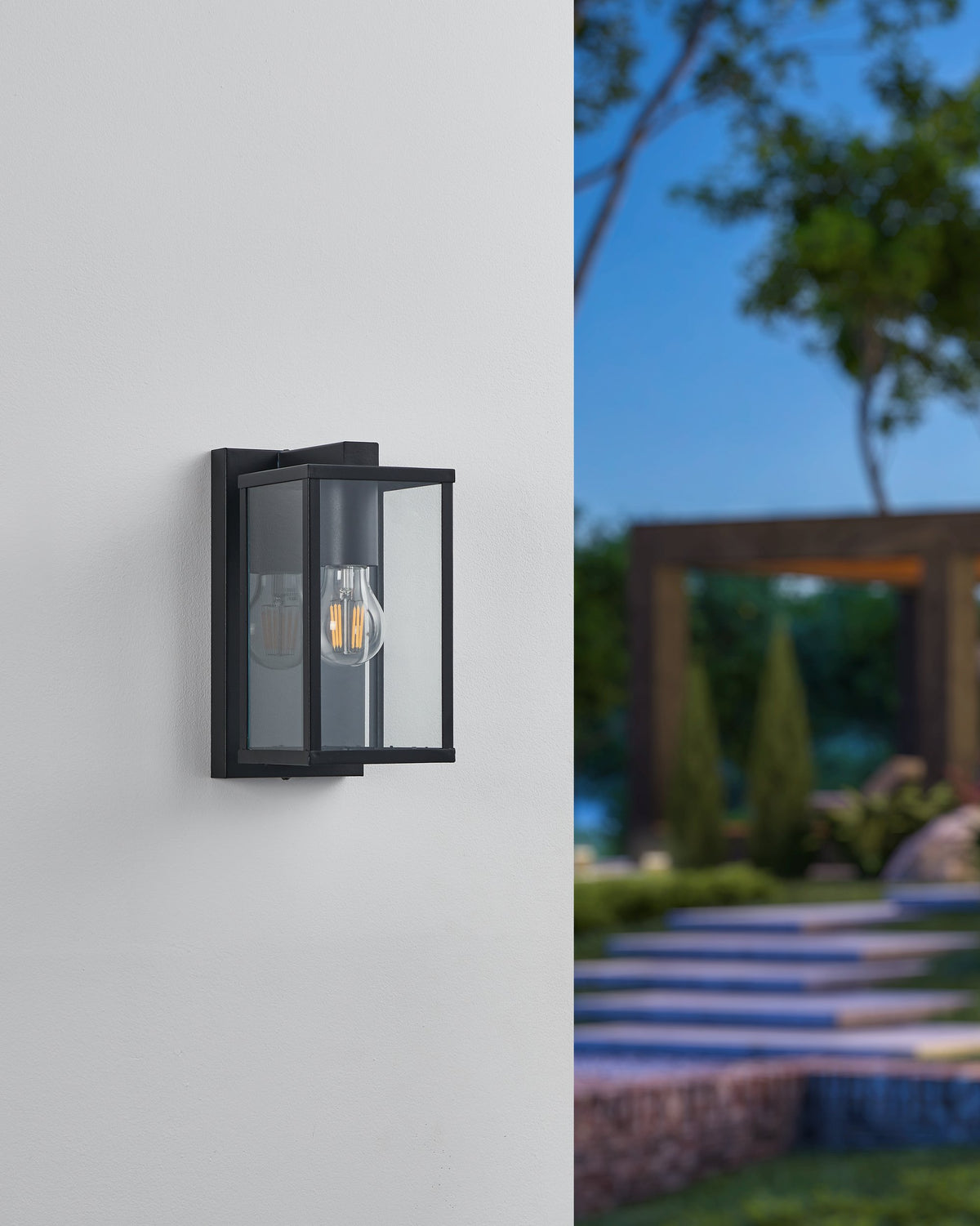 Même applique murale éteinte en journée, cadre noir et verre clair devant un jardin flou