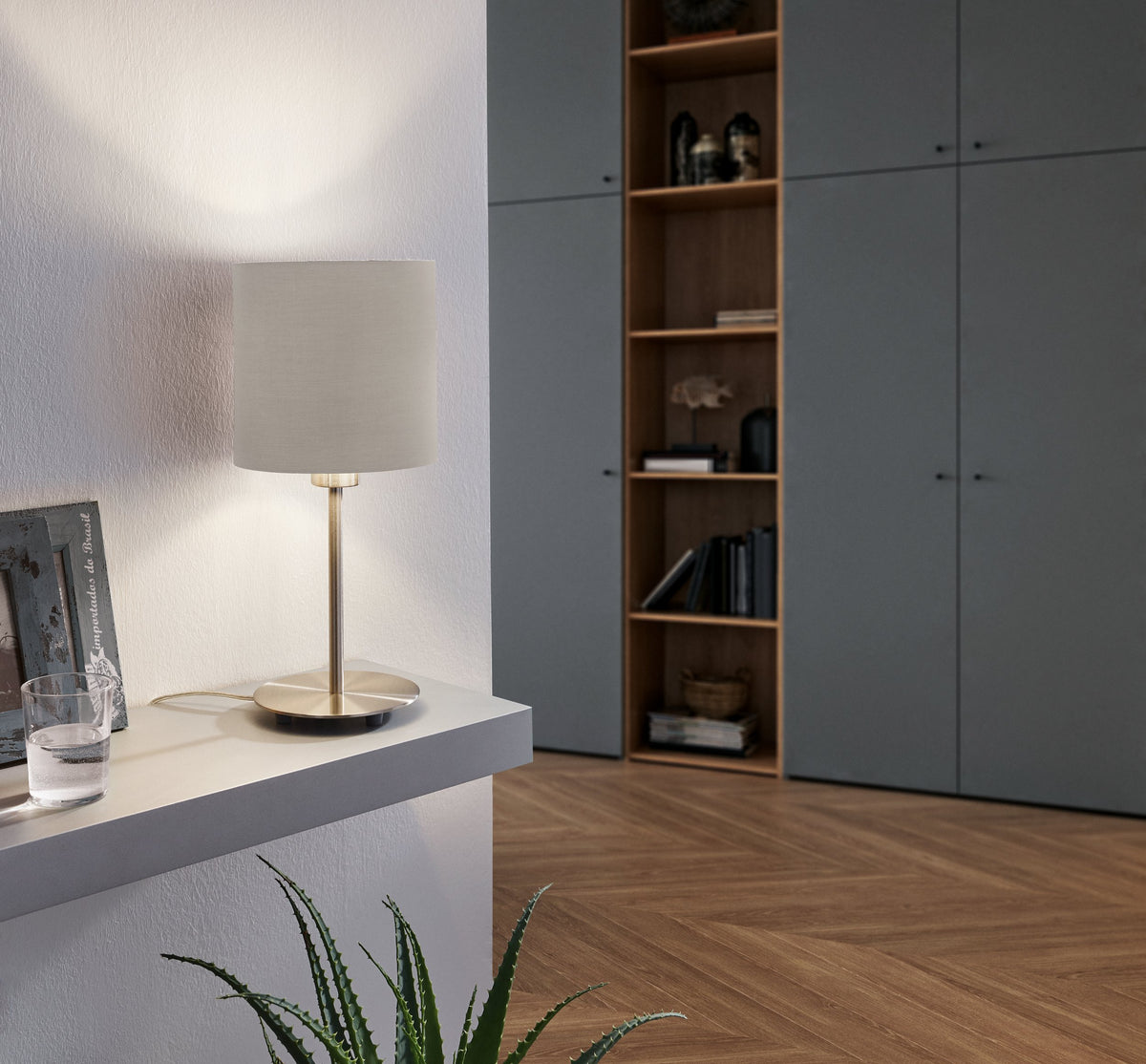 Lampe de bureau allumée sur une console devant une bibliothèque grise et un mur blanc