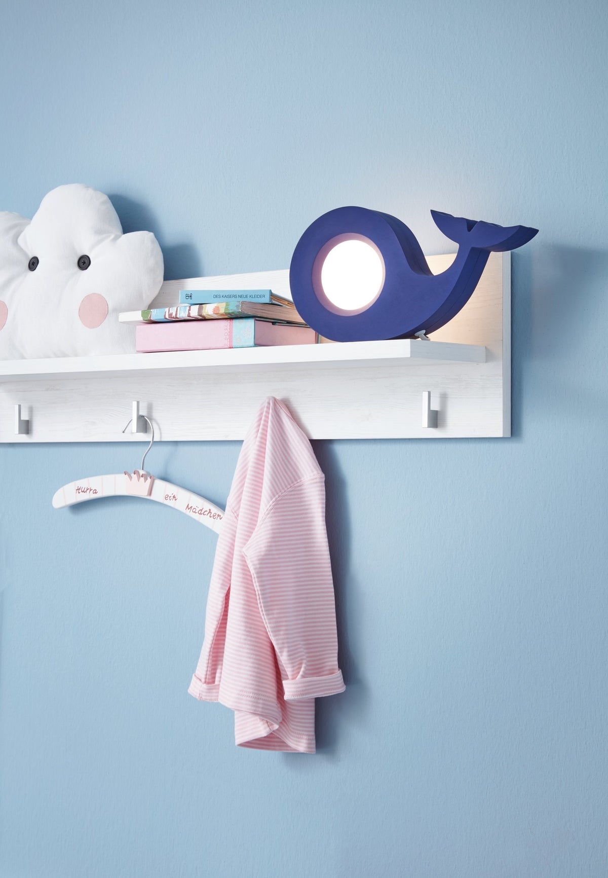 Lampe baleine bleue posée sur étagère blanche, près de livres et d'un vêtement rose.