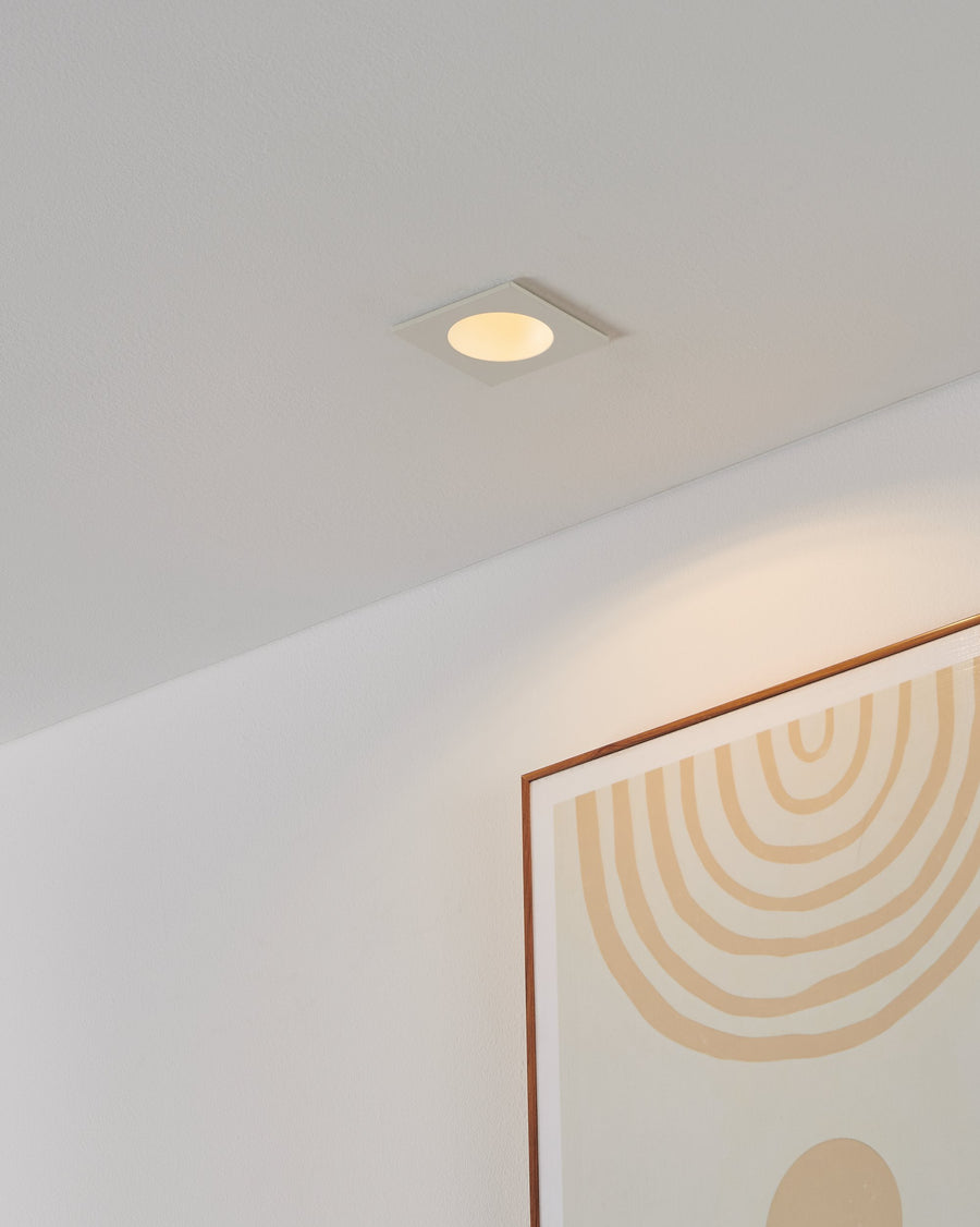 Spot encastré au plafond blanc, allumé, éclairant un mur et un cadre décoratif.