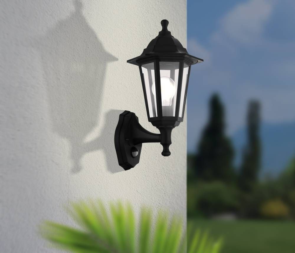 Applique extérieure noire installée sur un mur blanc, ombre portée et plantes floues