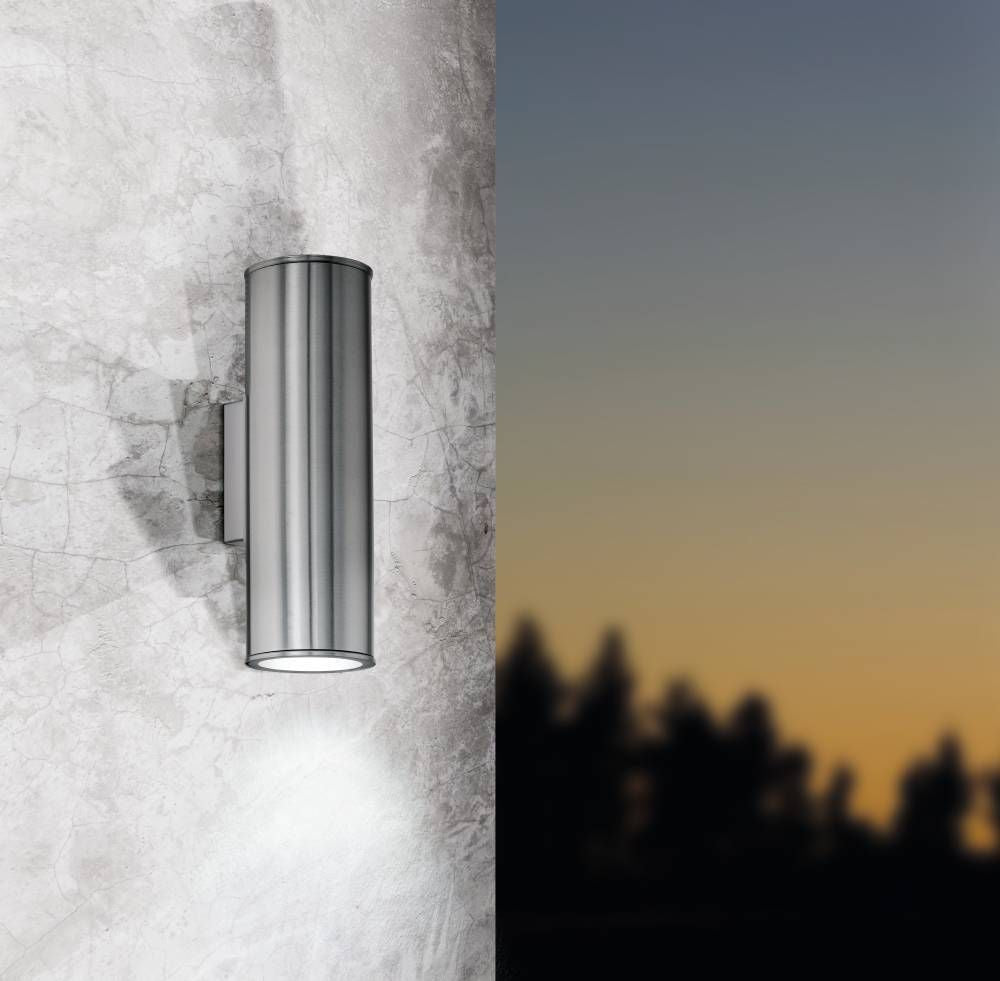 Applique murale cylindrique en inox allumée sur un mur extérieur, crépuscule en arrière-plan