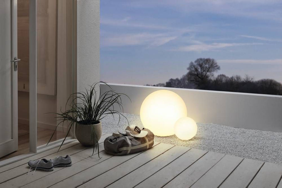 Deux sphères lumineuses blanches éclairent une terrasse en bois au crépuscule, plante et sac.